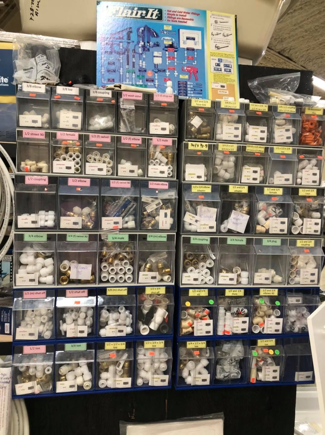 A wall display at a hardware store showing various plumbing fittings in clear plastic bins, labeled with pink price tags.