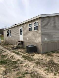 A single-story, beige mobile home with a white door, steps, and an air conditioning unit on a dirt lot.