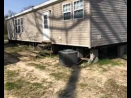 A single-story beige mobile home with visible undercarriage, windows, and an AC unit sits on a grassy lot.