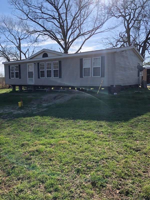 A light gray mobile home raised on cinder blocks on a grassy lot. Trees in the background.