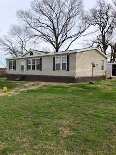 Tan mobile home with black shutters and brick skirting, set on a grassy lot with a large tree in the background.