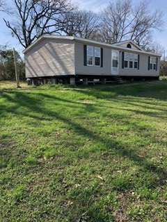 A light gray mobile home with black shutters is elevated on concrete blocks, in a grassy yard.
