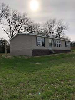 A light gray mobile home with black shutters sits on a grassy lawn under a bright sun. Trees line the background.