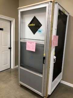 A tall, aluminum-framed booth with a screen door, a side door, and a sign, standing indoors near a closed white door.