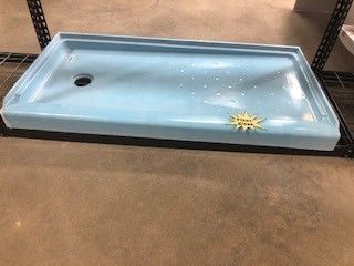Light blue rectangular shower base with drain, sitting on a black shelf in a warehouse setting.