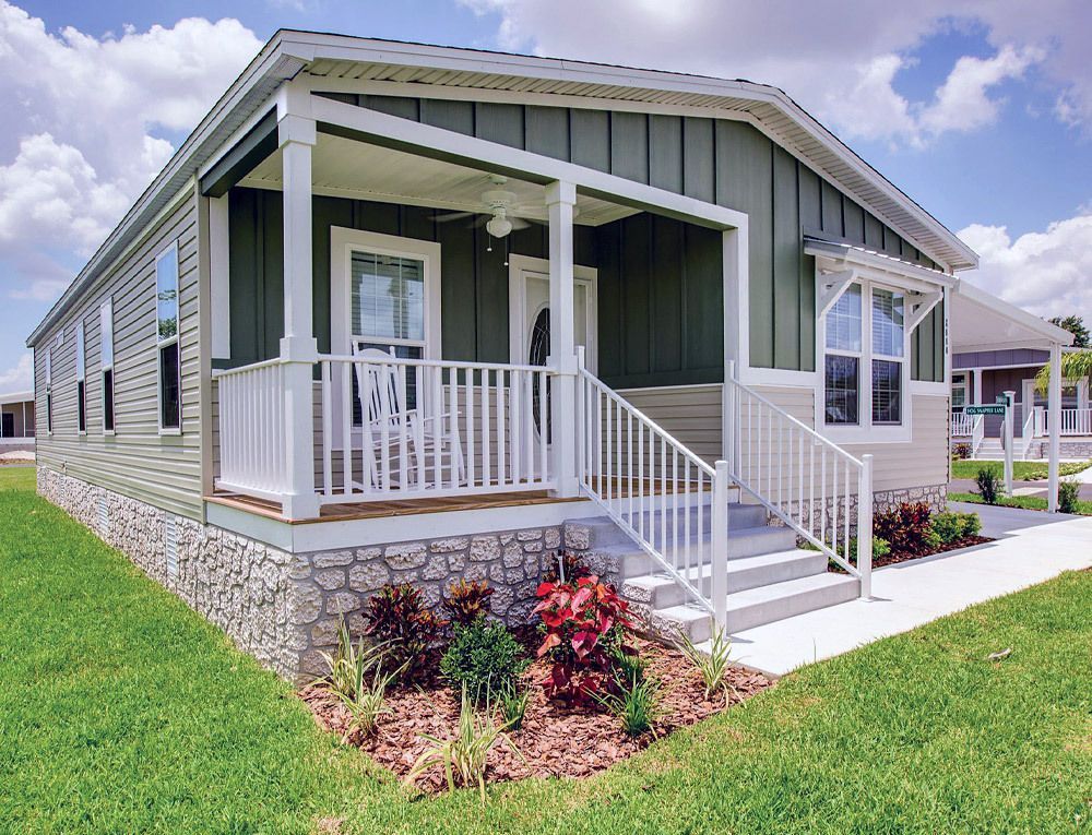 Green and white house with a covered porch, steps, and stone facade, set on a lawn with landscaping under a cloudy sky.