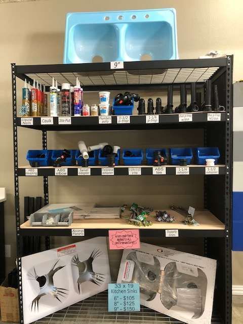 A metal shelf displays various plumbing supplies and two kitchen sinks. The top shelf features a light blue sink, with smaller items below.