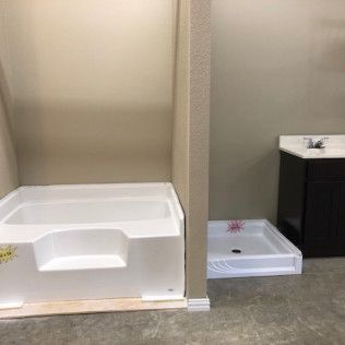 Bathroom fixtures on display: a white tub and a shower base sit next to a vanity with a sink. Light tan walls and a concrete floor.