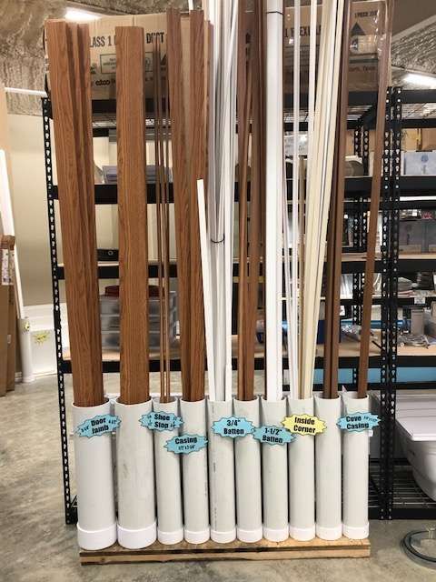 Wooden trim pieces of various colors and shapes are stored vertically in white tubes, labeled and arranged on a wooden base.