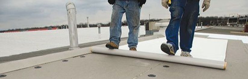 Two men are walking on a roof with a roll of roofing material.