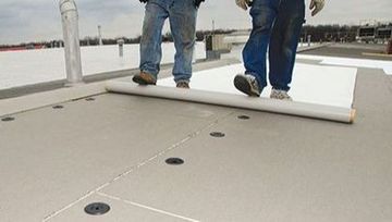 Two men are walking on a roof with a roll of roofing material.