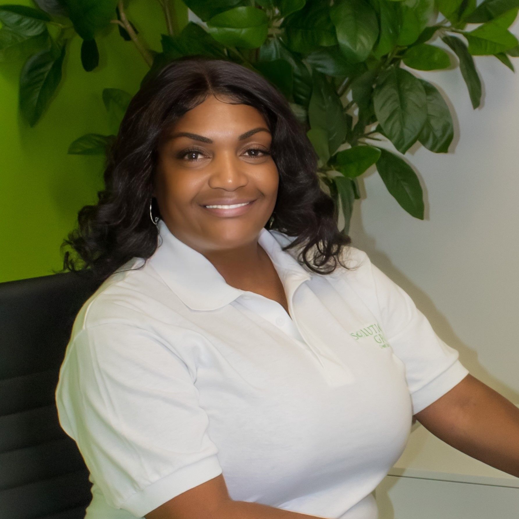 Woman in white polo smiling, seated near green plant, likely in an office setting.