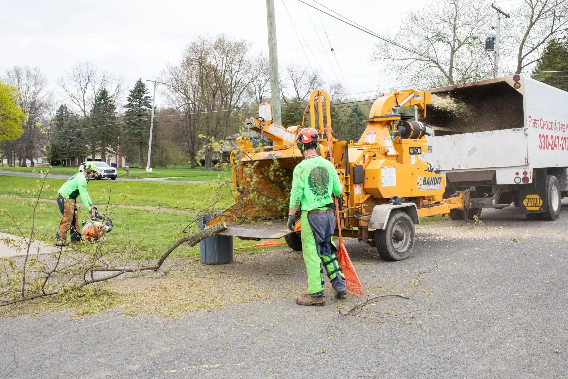 Two workers chipping tree branches into a truck with a wood chipper on a roadside.
