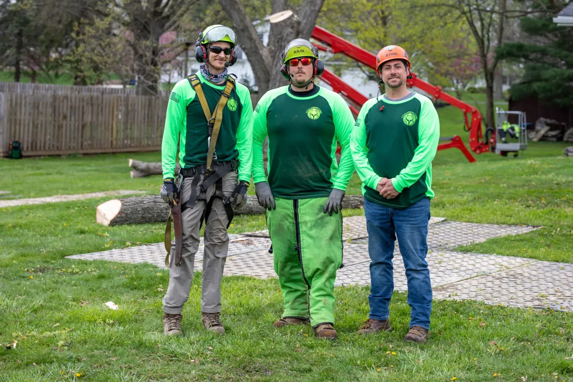 Three tree service workers posing outdoors near a felled tree and machinery, wearing green shirts and safety gear.