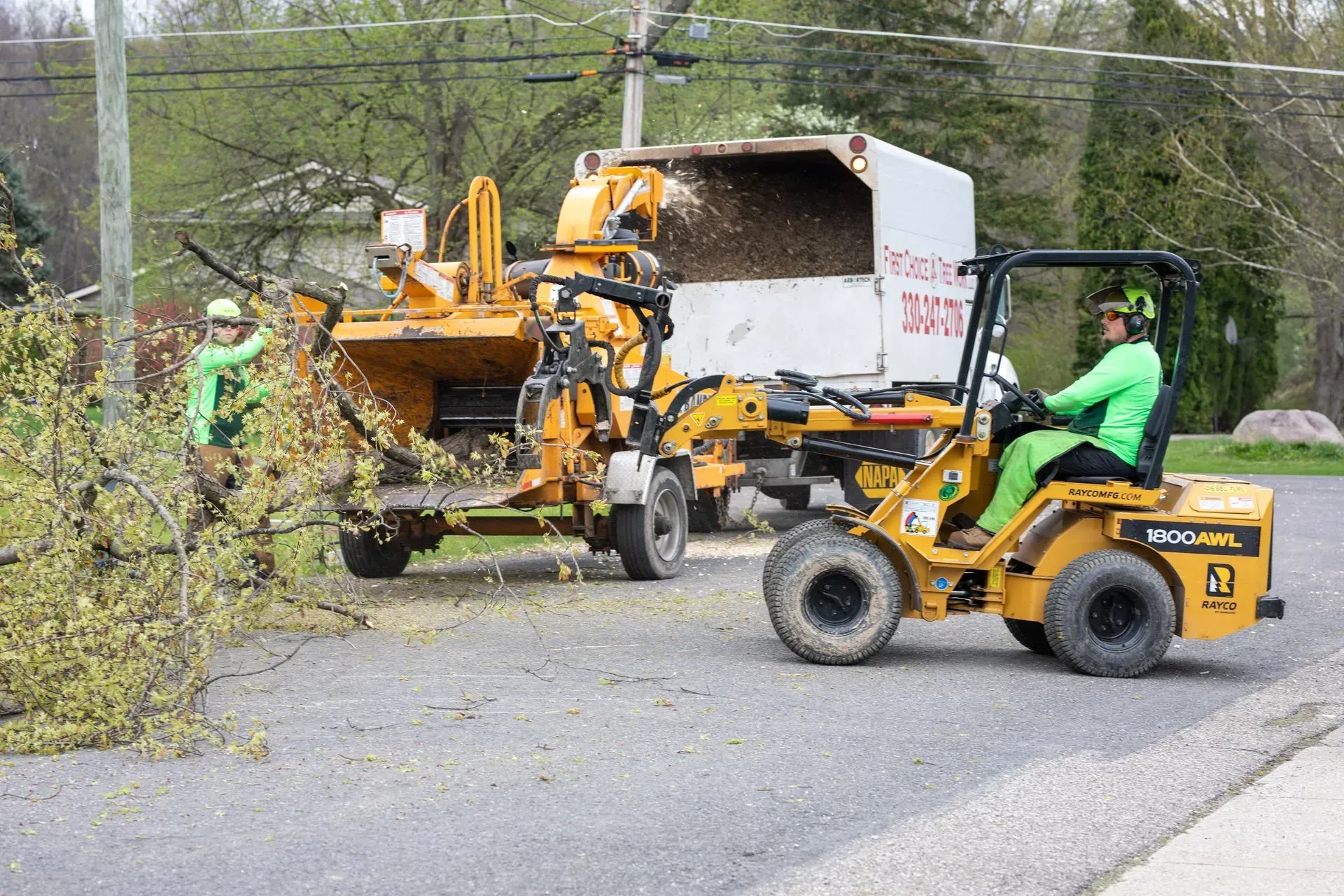 Two people use machinery to chip branches beside a road. One operates a loader, the other feeds branches into the chipper.