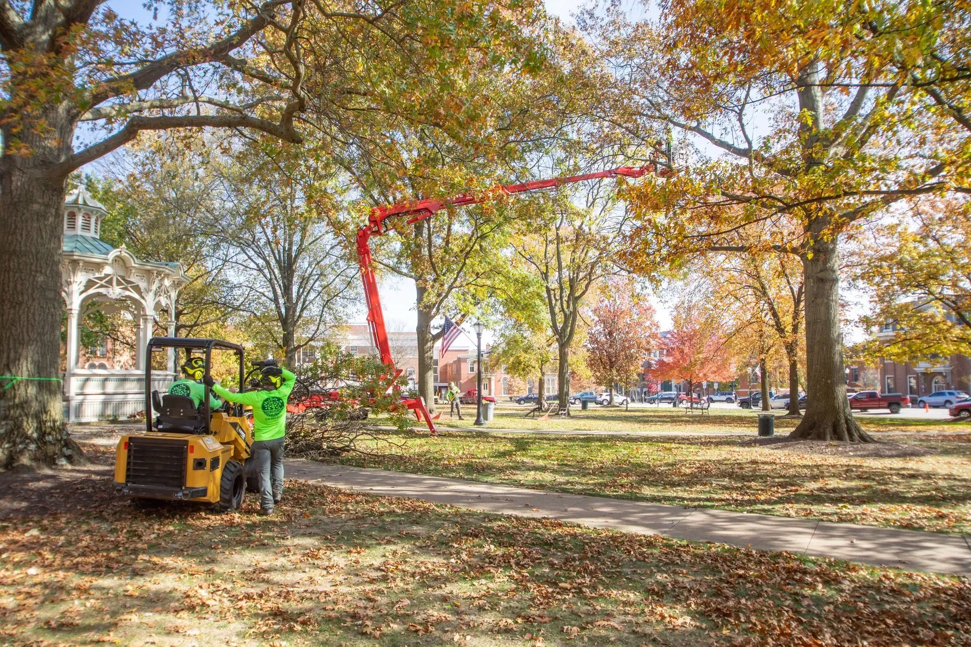 Workers trim tree branches with a lift in a park with fall foliage.