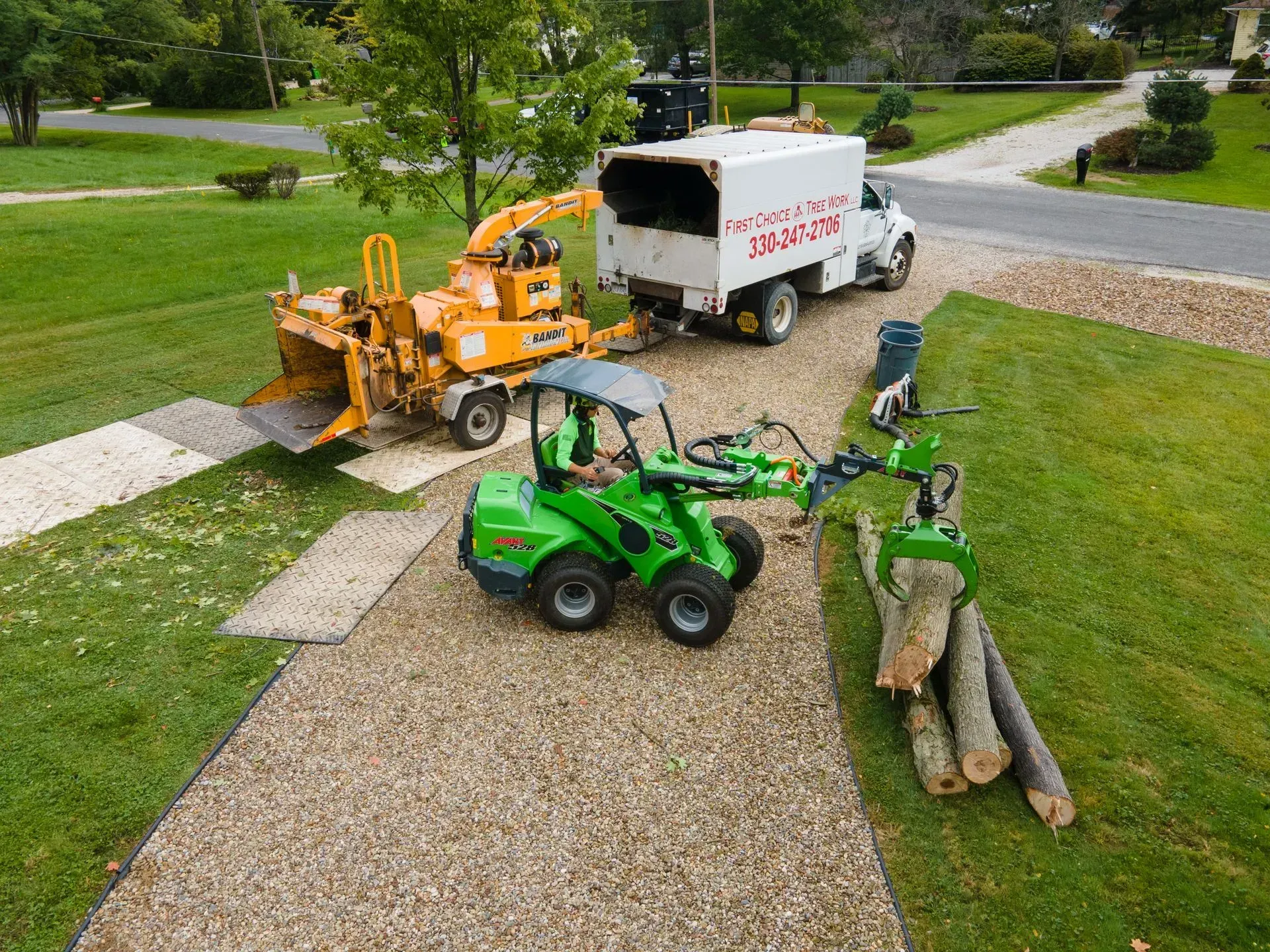 A tree service: Chipper, truck, and small green loader removing tree debris from a gravel driveway.
