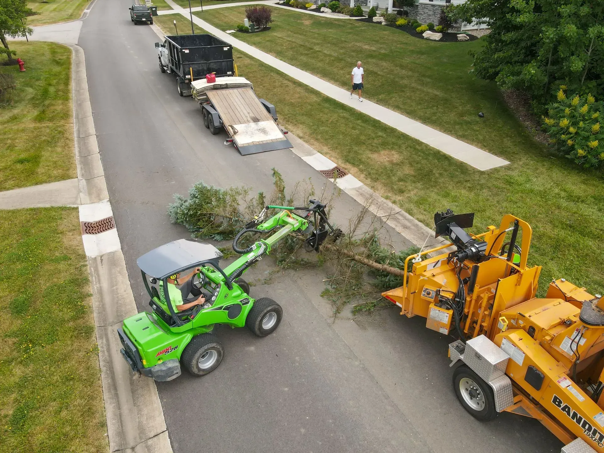 A green machine feeding tree branches to a wood chipper on a suburban street. A truck and trailer are nearby.