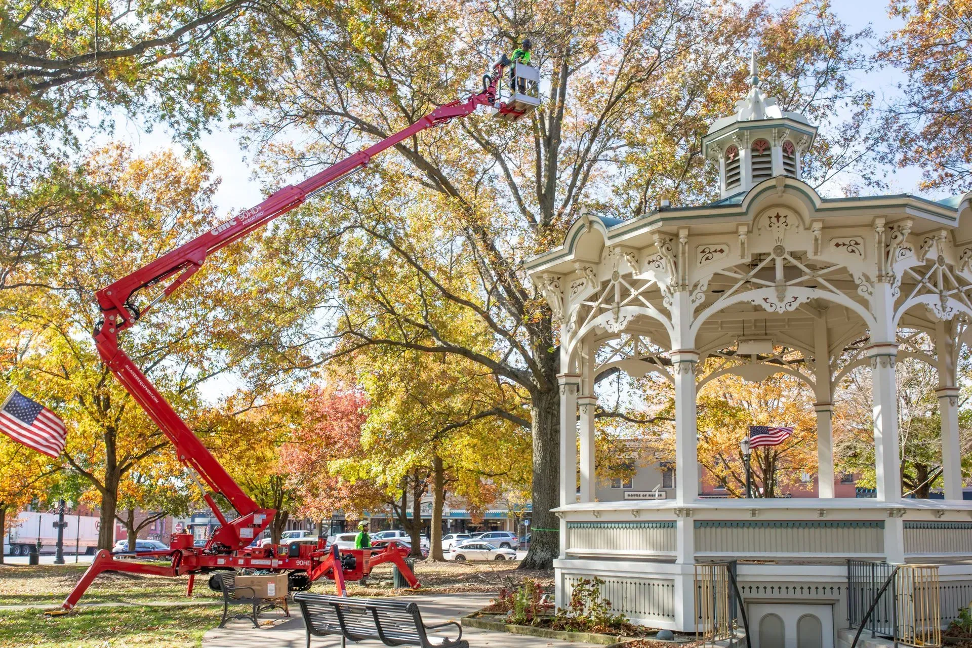 Red lift truck with a worker trimming tree branches near a white gazebo in a park, autumn setting.