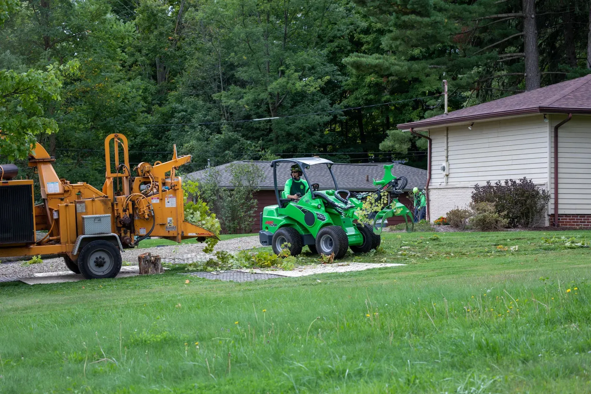 A worker operates a green mini-excavator to clear debris near a beige house and wood chipper.
