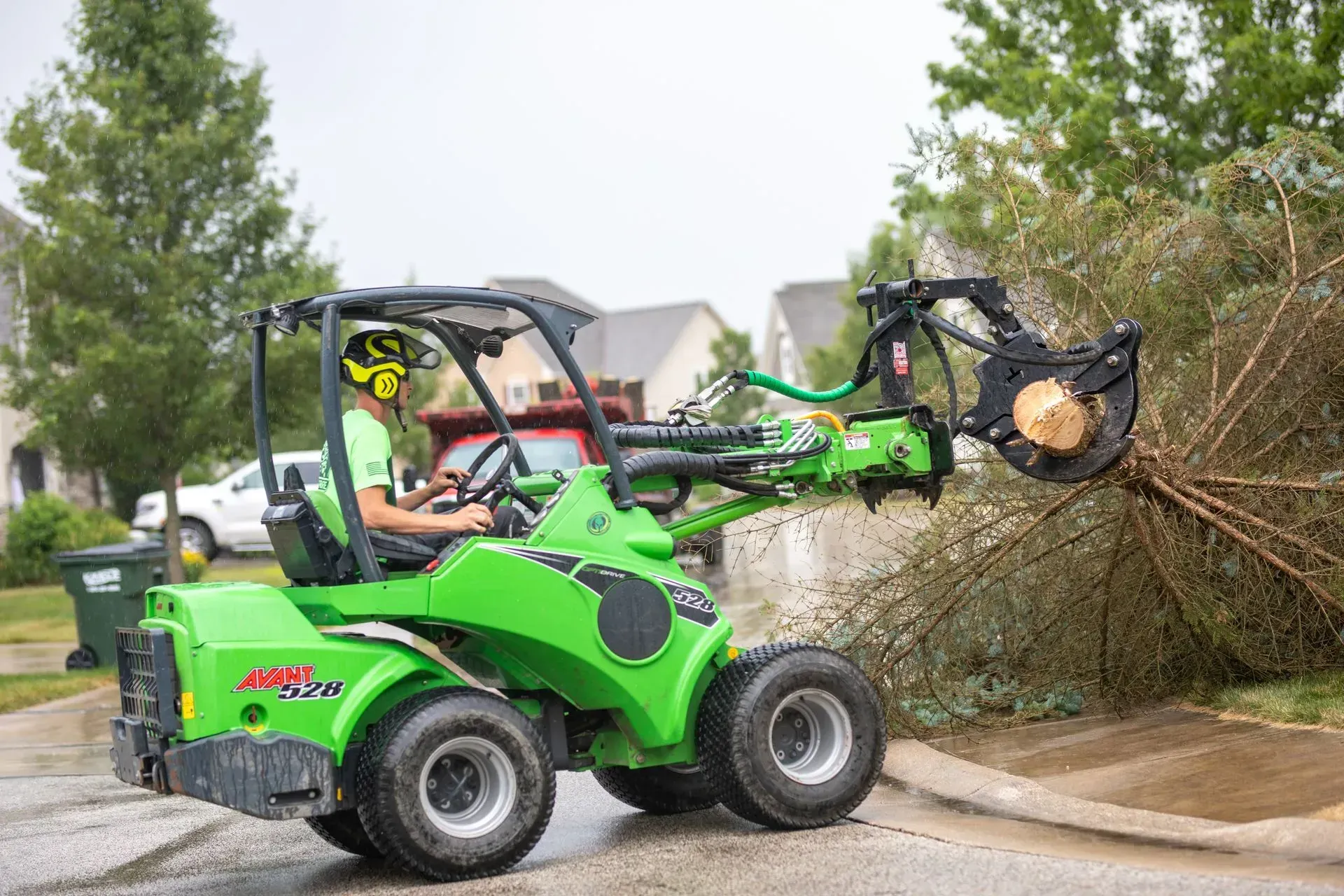 Green mini-loader with operator trimming tree branches on a street.