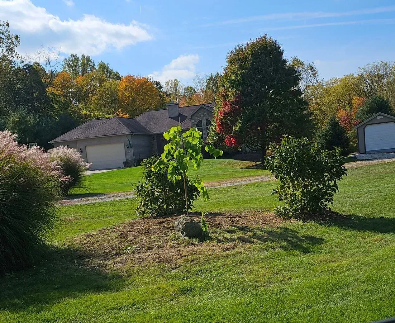 House with two garages set in a grassy yard, surrounded by trees with autumn foliage under a blue sky.