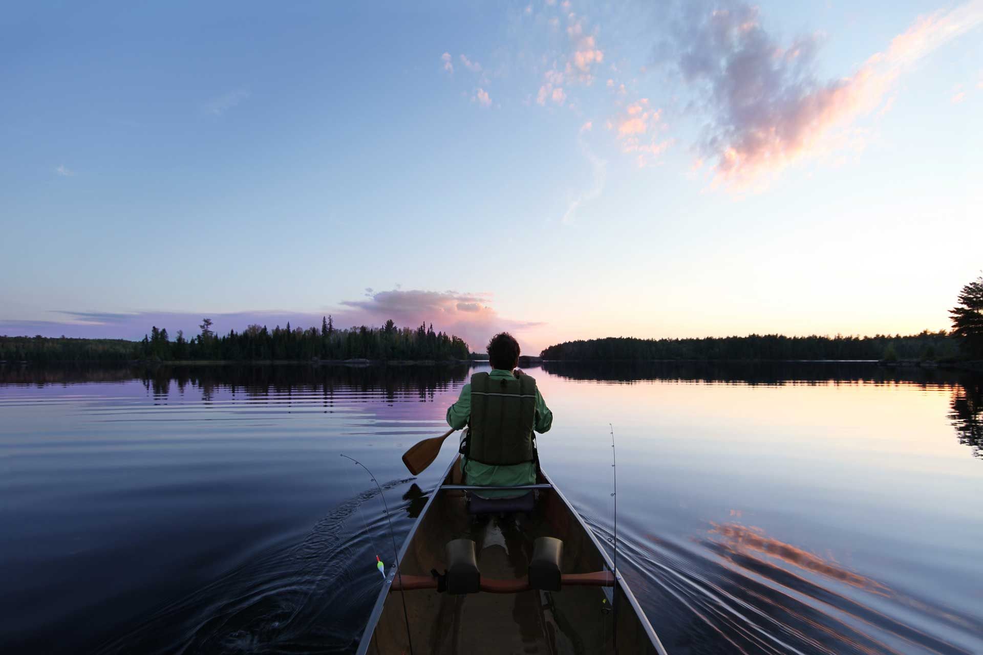 Person canoes on calm lake at sunset; trees line the shore, sky has pink and blue hues.