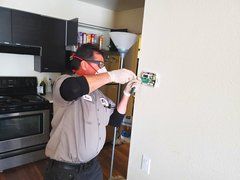 A man wearing a mask and gloves is working on a thermostat in a kitchen.