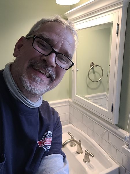 A man wearing glasses is smiling in front of a bathroom sink