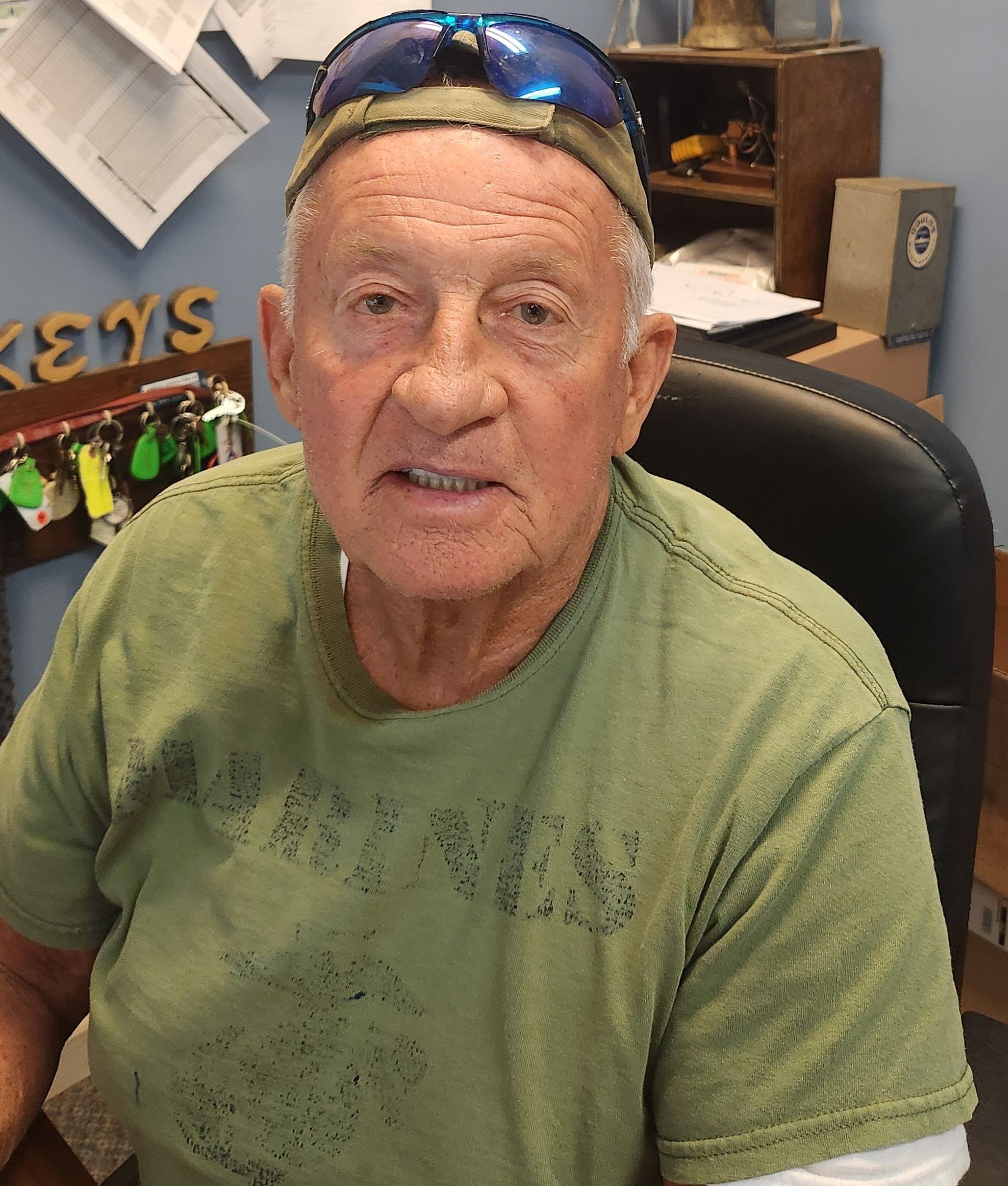 An elderly man with light skin, wearing a green shirt and blue sunglasses on his head, sits at a desk.