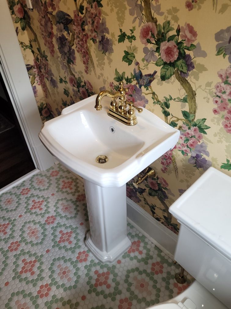 A white pedestal sink with gold fixtures stands against floral wallpaper, next to a white toilet.