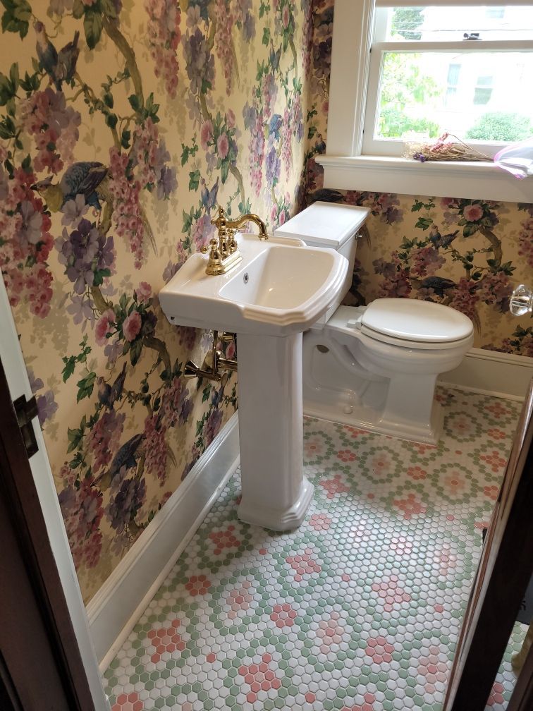 Bathroom with floral wallpaper, a sink with gold fixtures, and a white toilet atop of pink and green floral floor tiles.