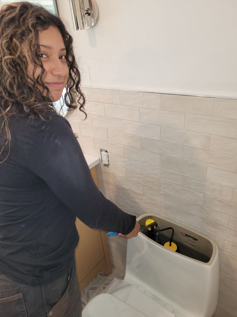 Woman working on a toilet in a bathroom. She is smiling while holding a blue tool inside the tank.