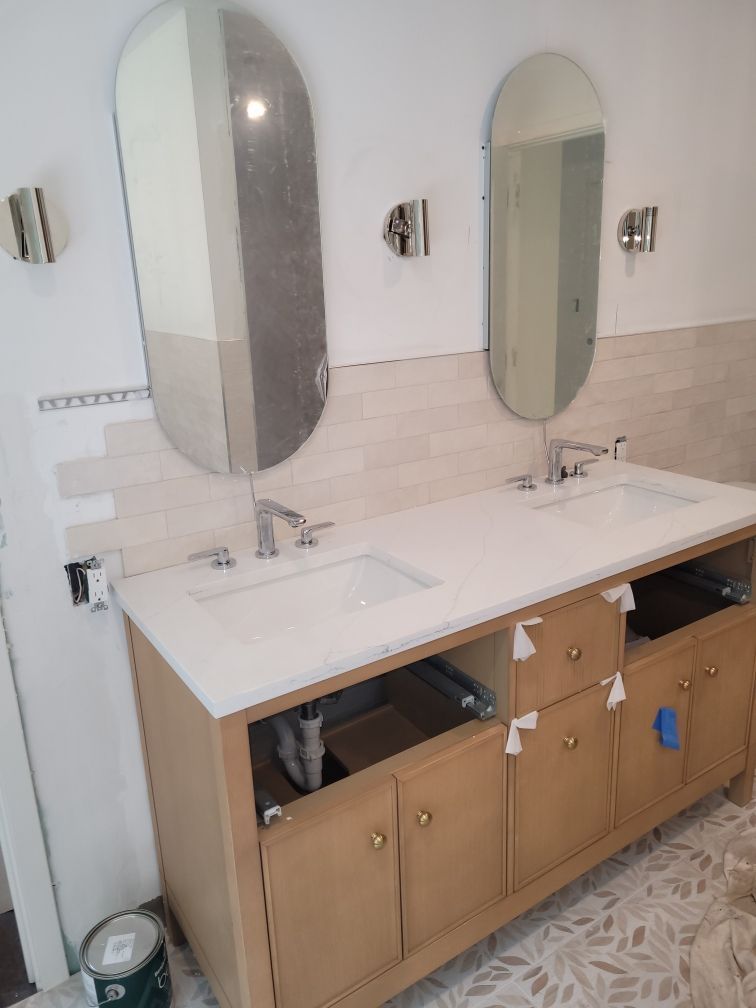 Bathroom vanity with light wood cabinets, two white sinks, oval mirrors, and chrome fixtures on light beige walls.