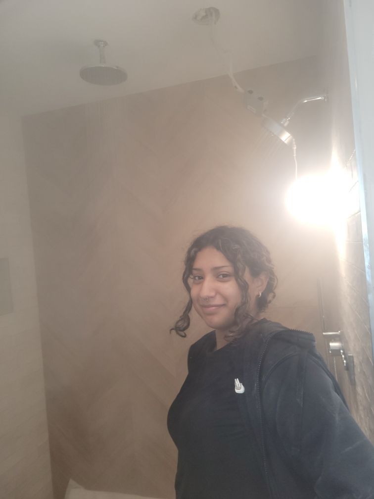 A woman with curly brown hair smiles in a bathroom, wearing a black outfit, near a bright light.