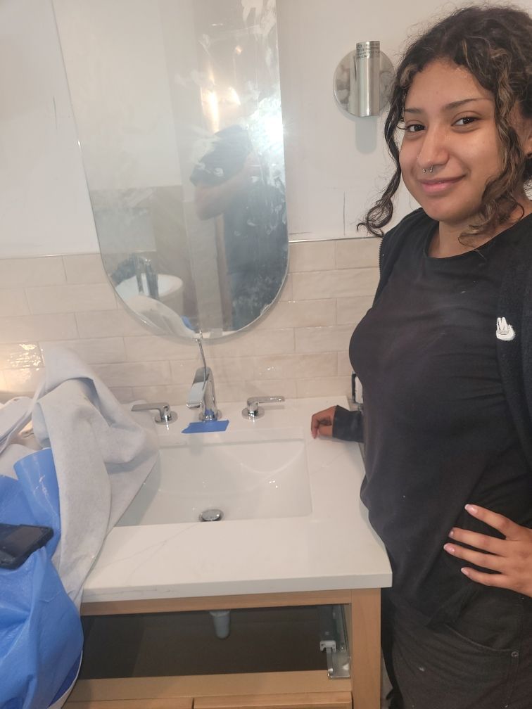Woman in front of a newly installed bathroom sink. She smiles with one hand on her hip. Beige tiles and a curved mirror are behind her.