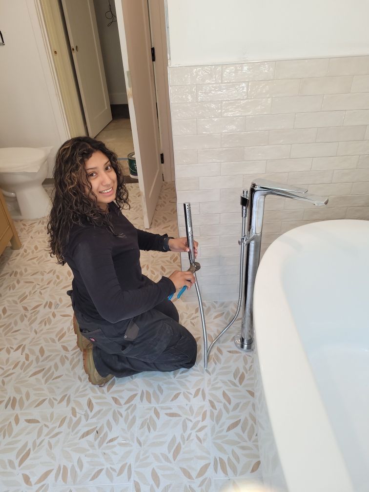 Woman kneels in a bathroom, assembling a chrome faucet near a tub. She smiles at the camera; the room has patterned tile and white walls.