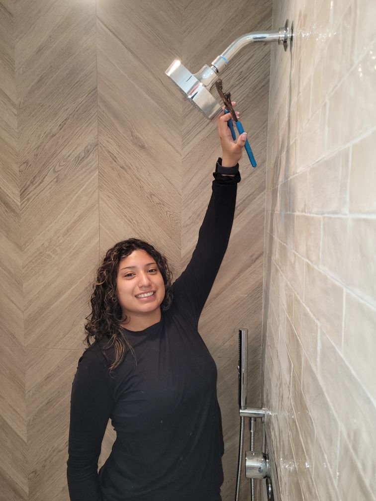 Woman in black top holding a wrench while working on a shower head in a tiled bathroom. She smiles at the camera.