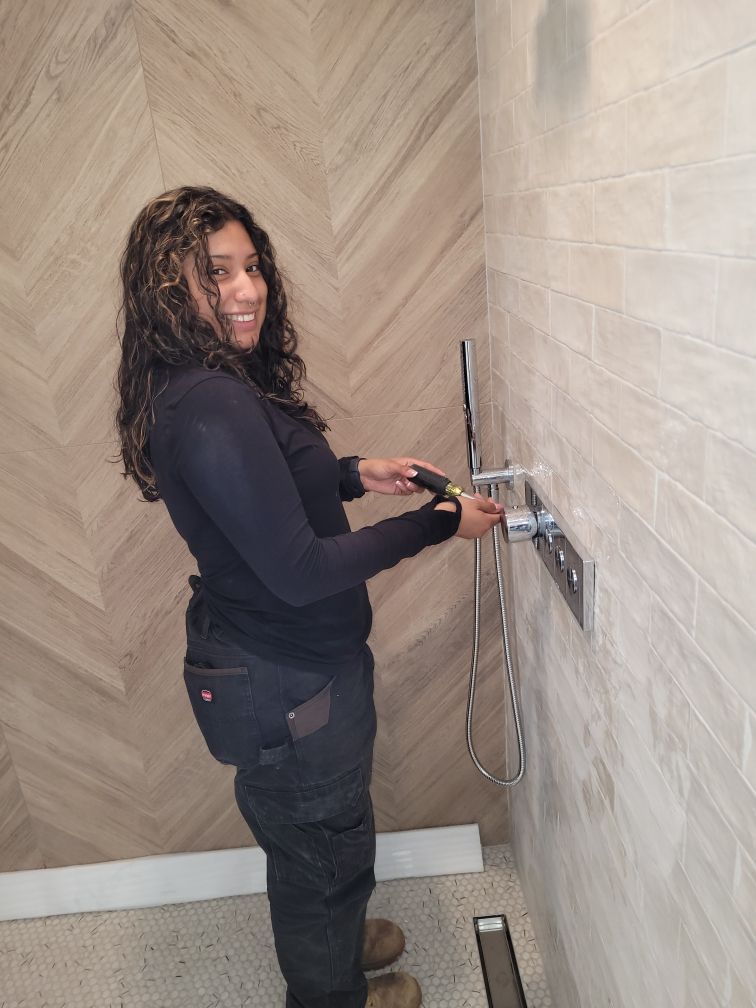 Woman in work clothes smiling while installing shower fixtures in a bathroom with tile walls.