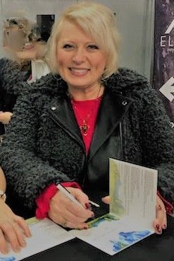 Woman with blonde hair smiles while signing a book. She wears a black jacket over a red shirt and sits at a table.