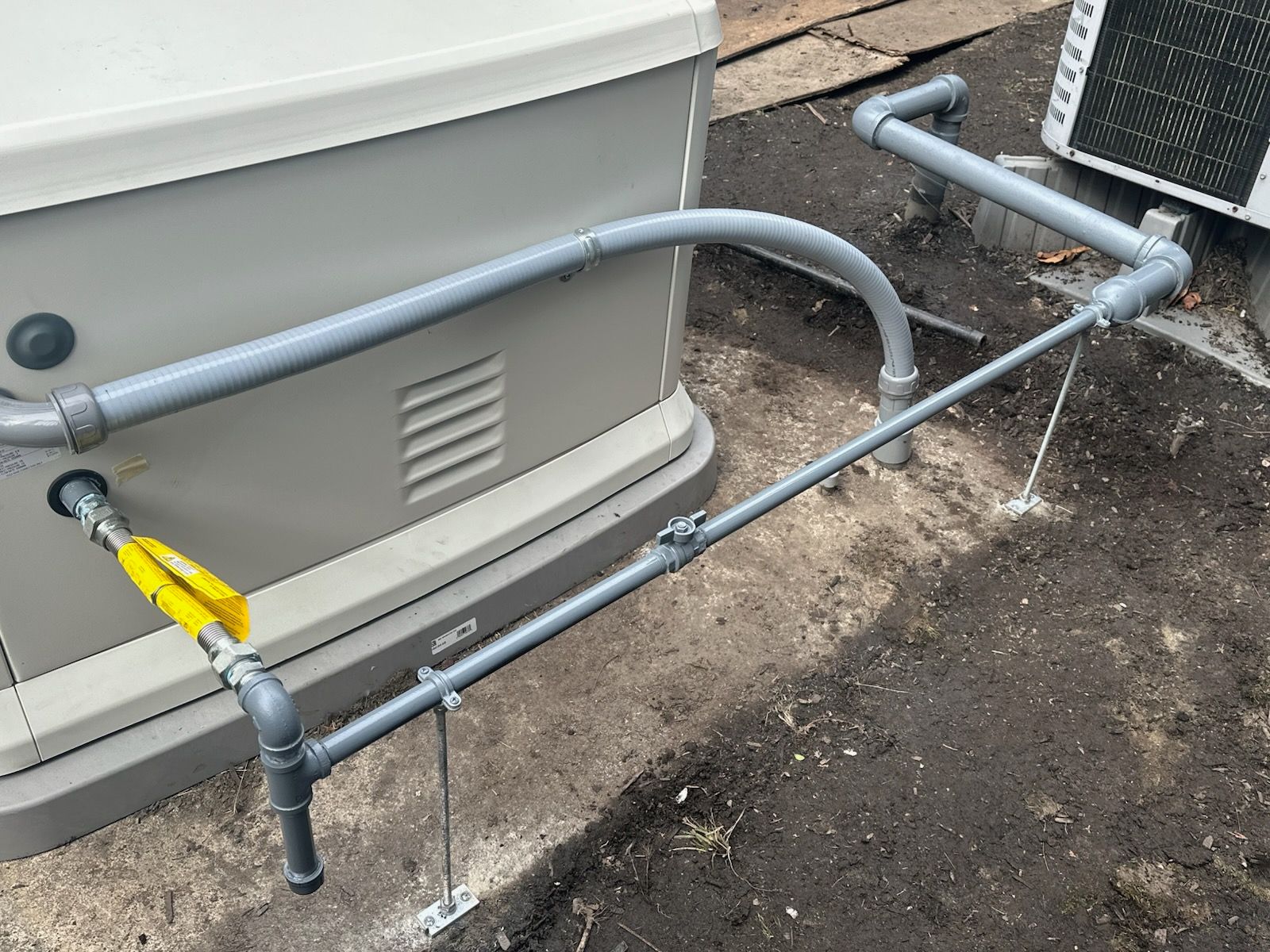 A gray generator with gas piping. The pipes are gray, supported by metal brackets and the yellow gas valve is open.