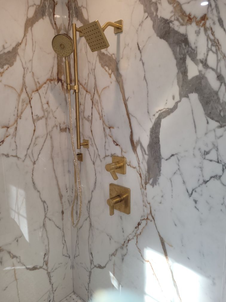 Gold shower fixtures against a white marble-veined wall.  The showerhead and hand-held sprayer are visible.