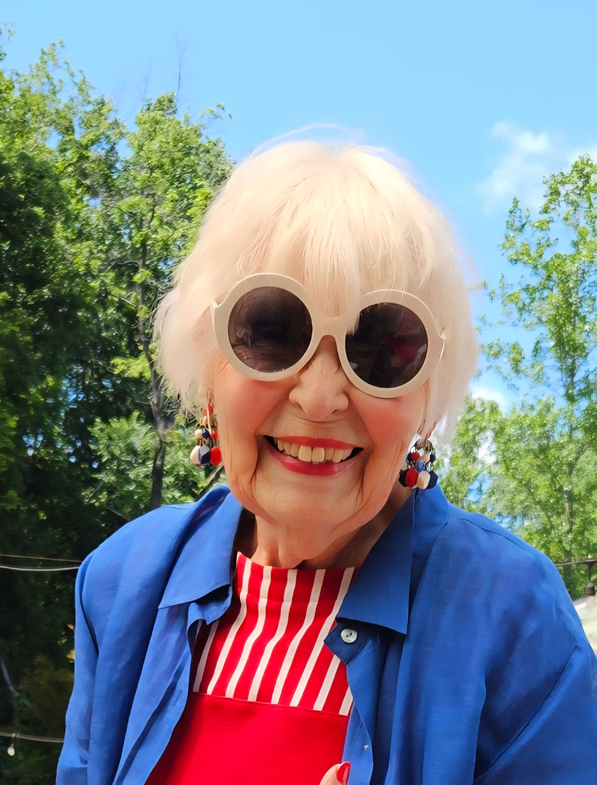 Woman with white hair wearing large white sunglasses, red and white striped shirt, blue jacket, and patriotic earrings smiles outside.