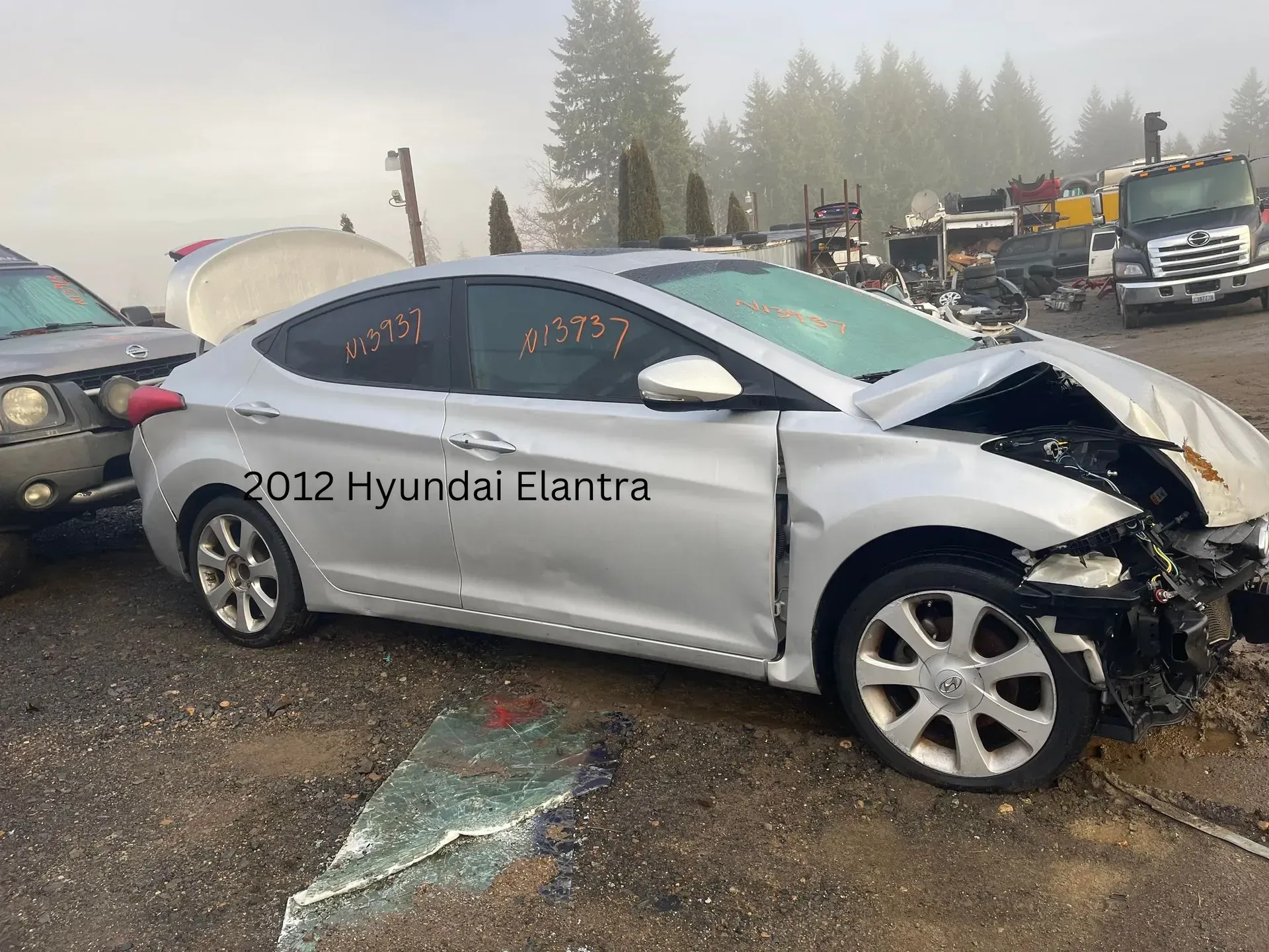Damaged silver 2012 Hyundai Elantra at a junkyard, hood crumpled, windshield broken.