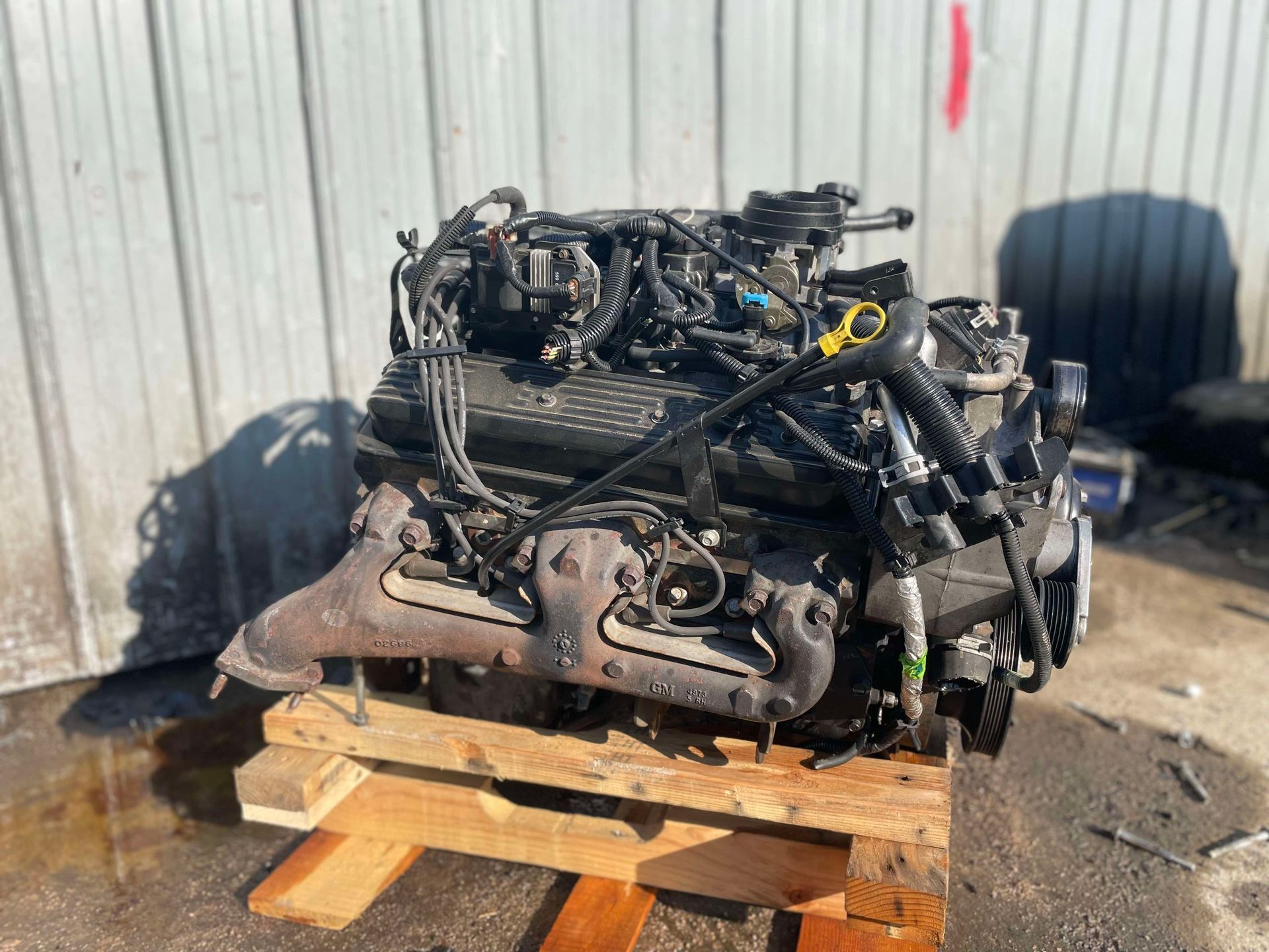 Engine on a wooden pallet; black and rusty metal with various wires and hoses.
