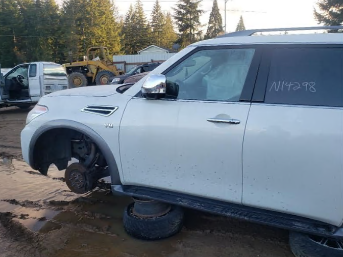 White SUV in a junkyard, missing a wheel. Muddy ground, other vehicles visible.