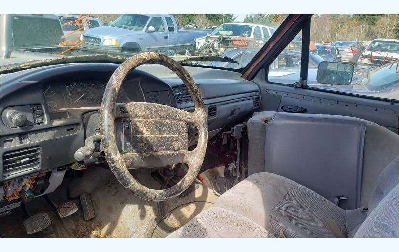 Dusty interior of a light-colored Ford truck, steering wheel and seats are dirty, parked outdoors with other vehicles.