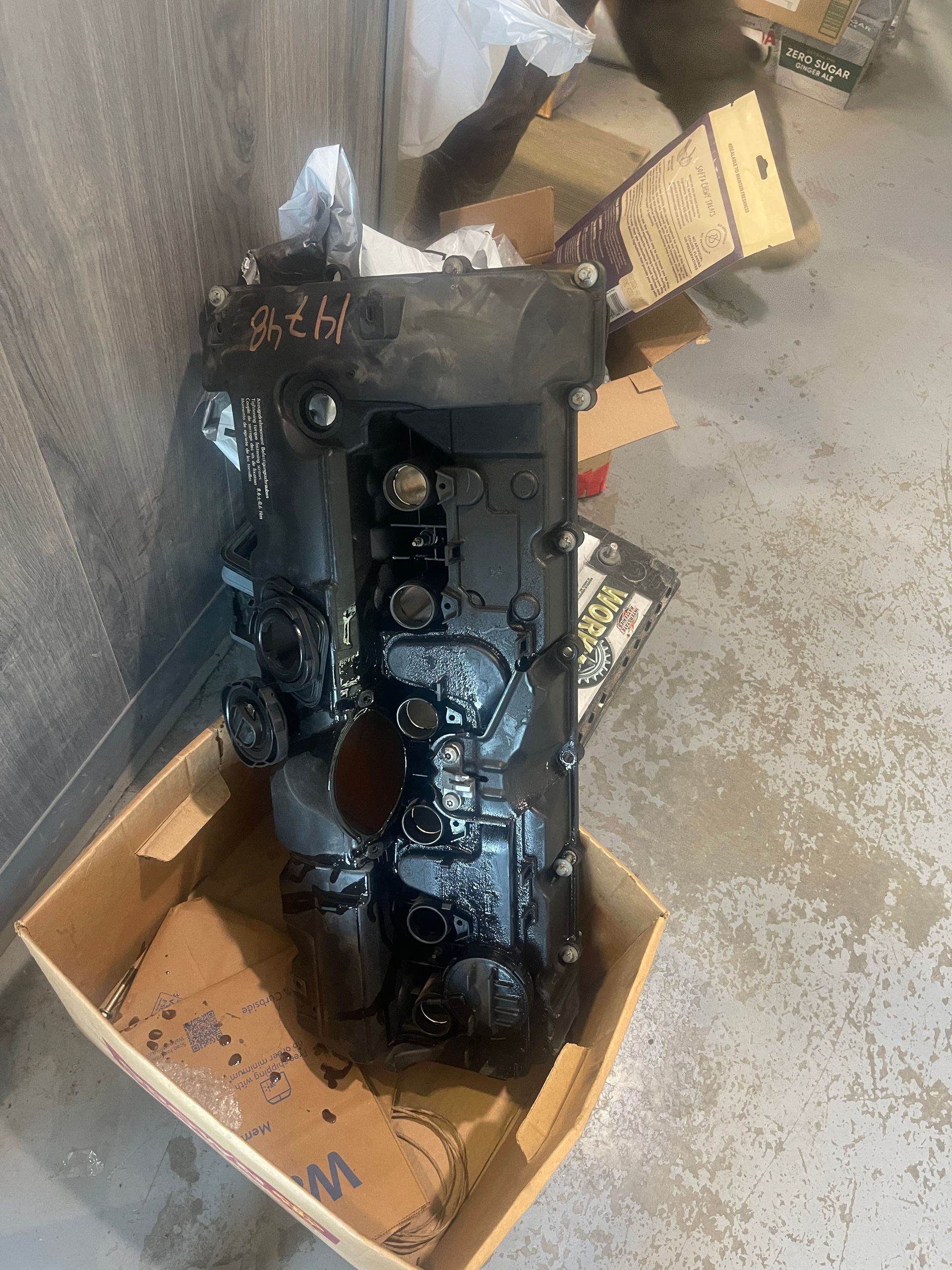 Black mechanical part covered in oil, in a cardboard box, indoors.
