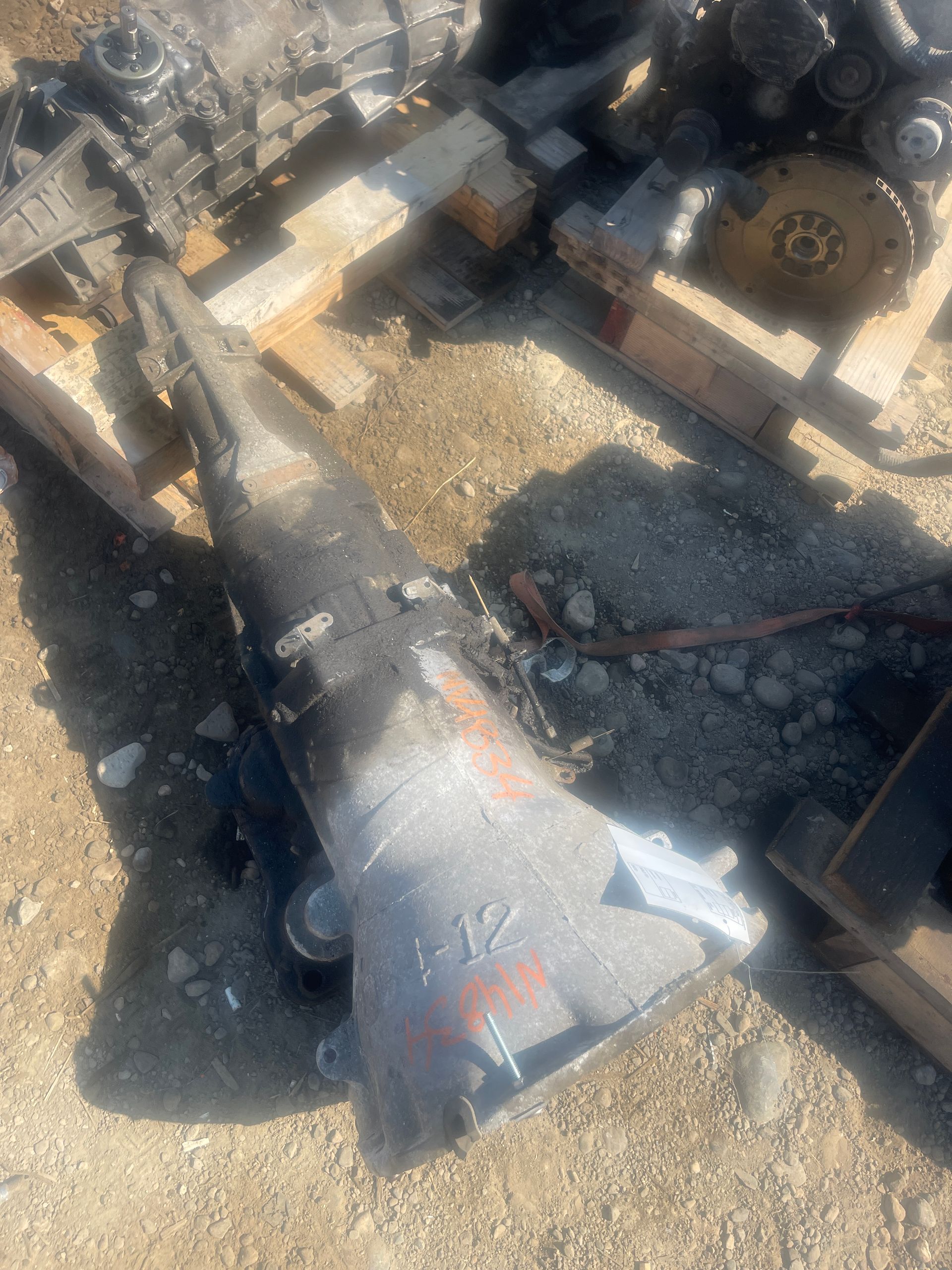 A used, gray automatic transmission lying on the ground with other auto parts in a salvage yard.