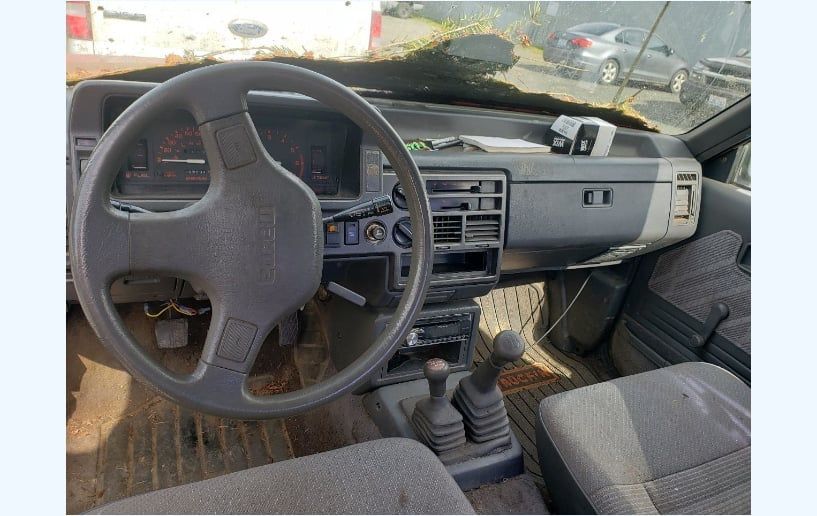 Interior of an old car, showing steering wheel, dashboard, seats, and gear shift. Gray and brown colors.